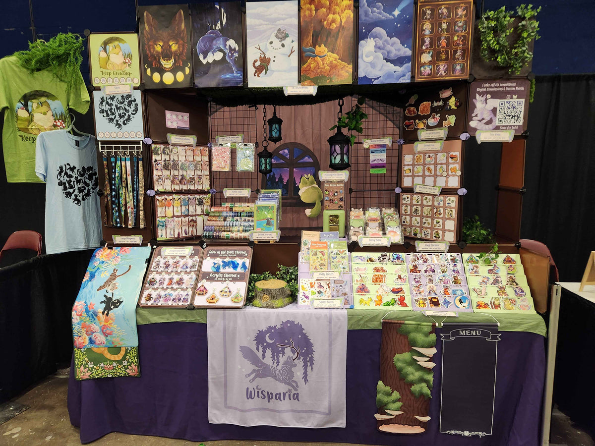 A market booth display for Wisparia, featuring a purple draped tablecloth, a central white cloth with a purple tree logo, various illustrated stickers, cards, and small goods arranged on tables and racks, with artwork hanging above.
